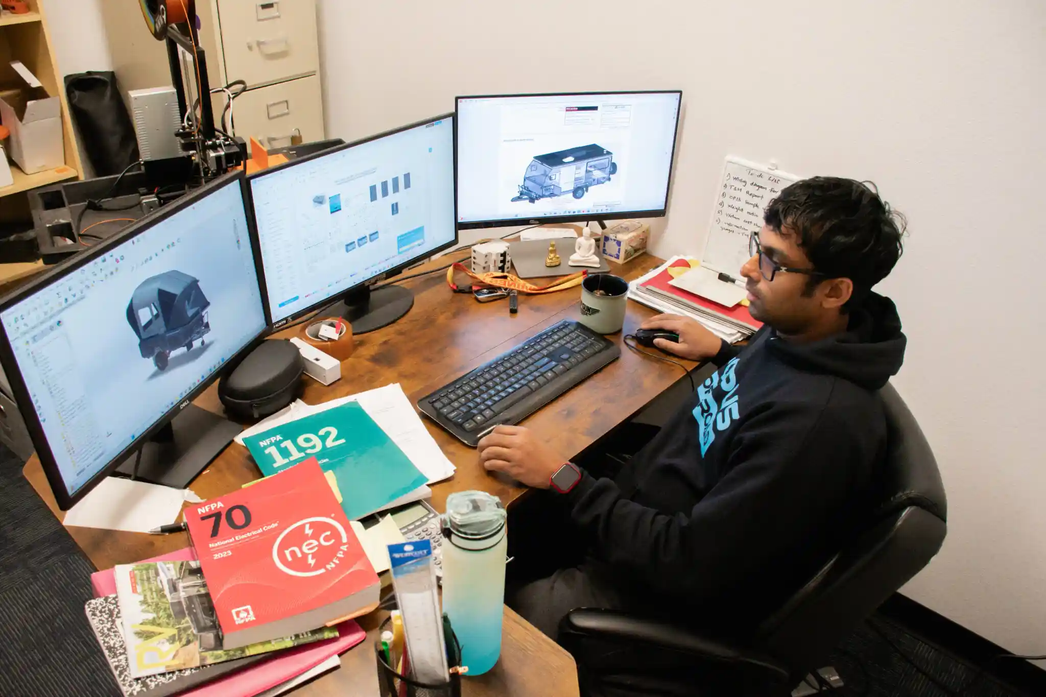 An OPUS engineer working on camper CAD drawings at a multi-monitor workstation, with NFPA code books on the desk